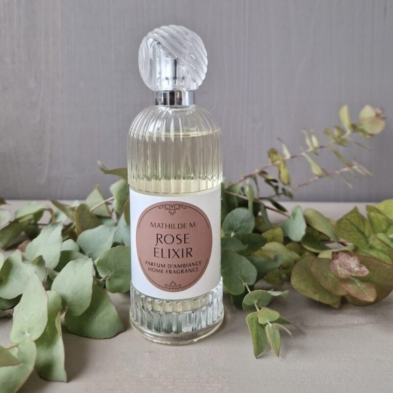 Parfumflakon Rose Elixir von Mathilde M, umgeben von grünen Blättern auf einem Holztisch.