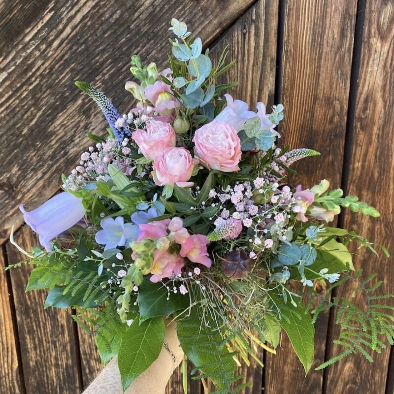 Bouquet aus rosa Rosen und lila Blumen, umgeben von grünem Laub, vor einer Holzwand. Romantische Blumendeko.