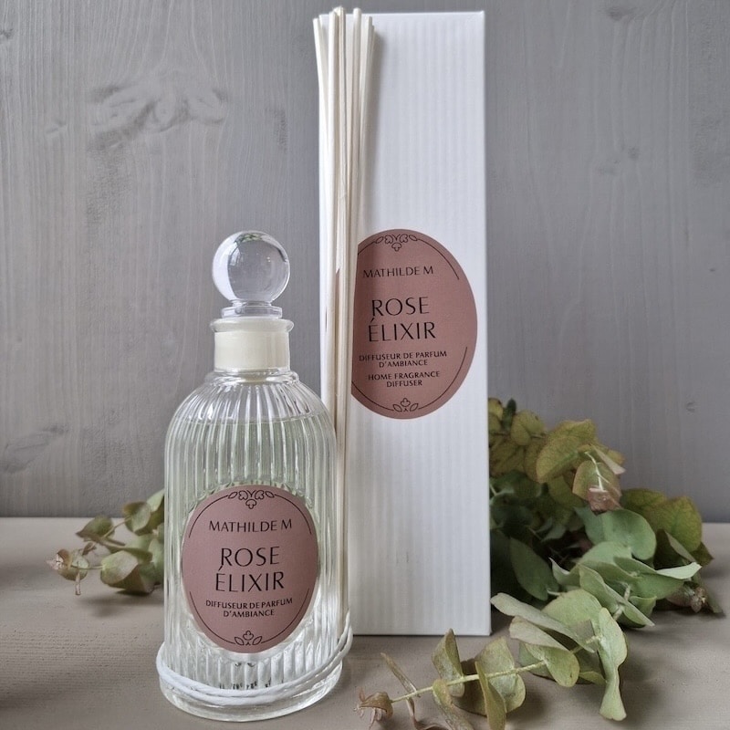 Mathilde M Rose Elixir Diffusor vor weißer Schachtel, umgeben von grünen Zweigen auf Holzoberfläche.
