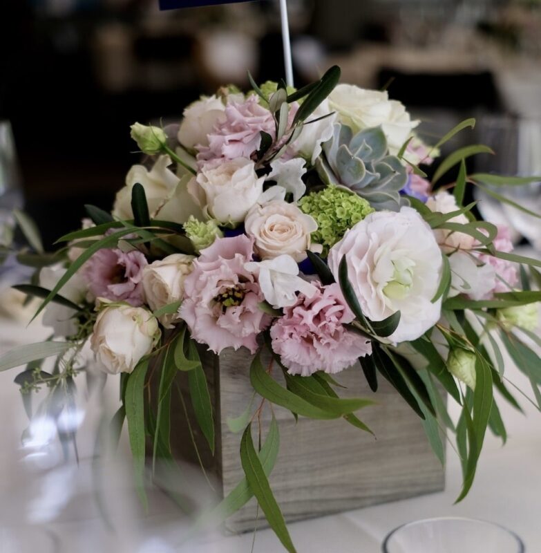 Blumengesteck aus rosa und weißen Blüten mit grünen Blättern in einer quadratischen Vase auf einem Tisch.