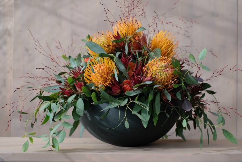 Blumenarrangement mit gelben Protea-Blüten und grünen Blättern in schwarzer Schale auf Holztisch.