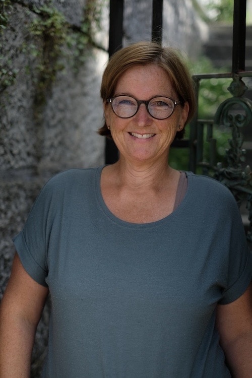 Frau mit Brille lächelt vor einer Steinmauer, sommerliches Ambiente.