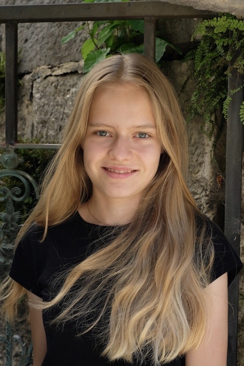 Junge Frau mit langen, blonden Haaren lächelt vor steiniger Wand und Pflanzen im Sommerlicht.