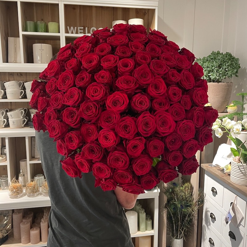 Großer Strauß aus roten Rosen in einem stilvollen Interieur mit Regalen und Dekorationen im Hintergrund.