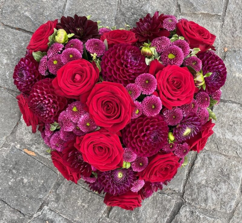 Herzförmiges Blumenarrangement mit roten Rosen und lila Chrysanthemen auf grauem Steinboden.
