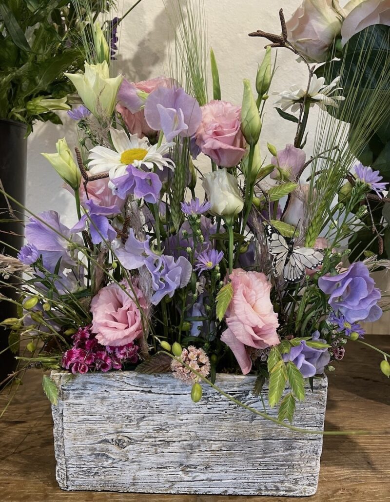 Bunter Blumenkasten mit Rosen, Margeriten und Schmetterling dekoriert, auf Holztisch arrangiert.
