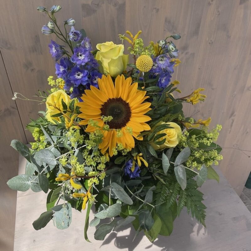 Bunter Blumenstrauß mit Sonnenblume, gelben Rosen, blauen Rittersporn und grüner Dekoration in natürlichem Licht.