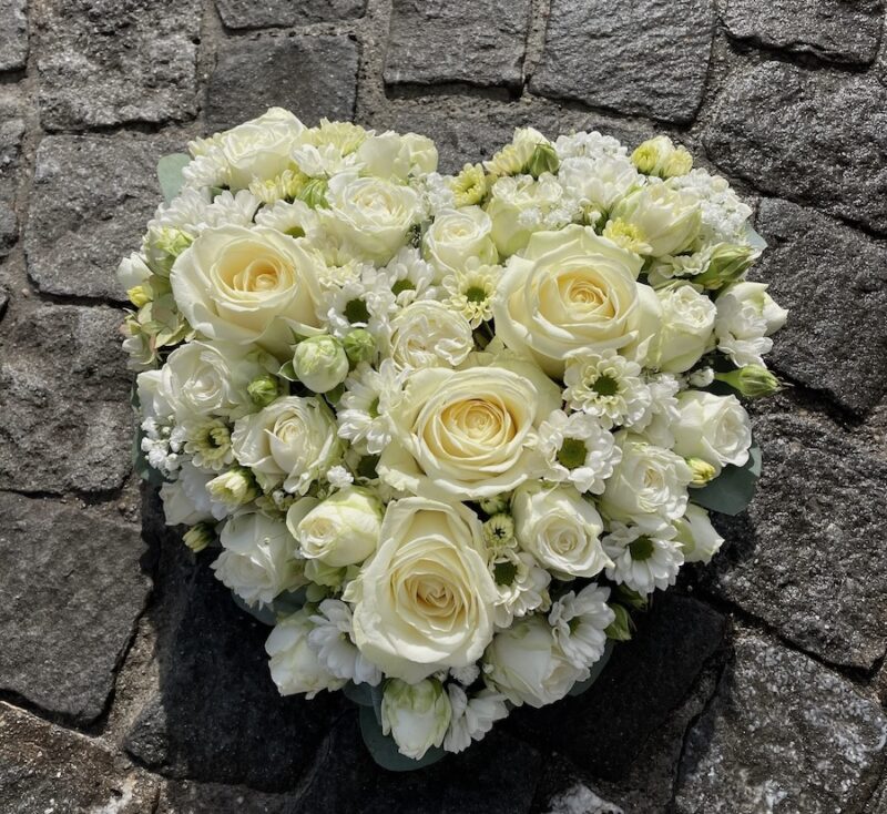 Herzförmiges Blumenarrangement aus weißen Rosen und Gänseblümchen auf Kopfsteinpflaster.