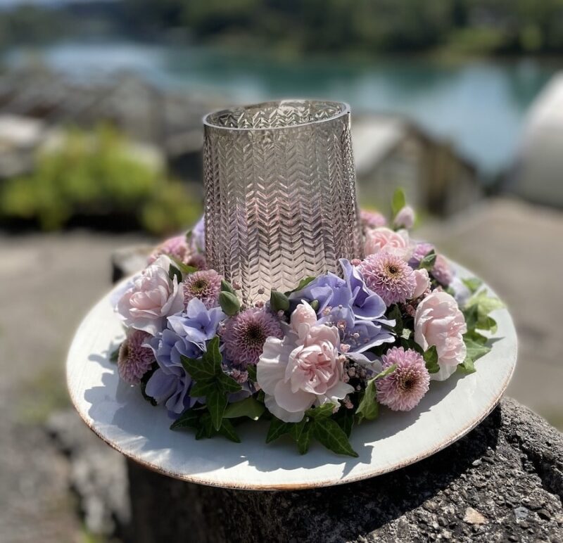 Blumengesteck mit rosa und lila Blumen umgibt einen Glaszylinder im Freien, dekorativ und elegant präsentiert.