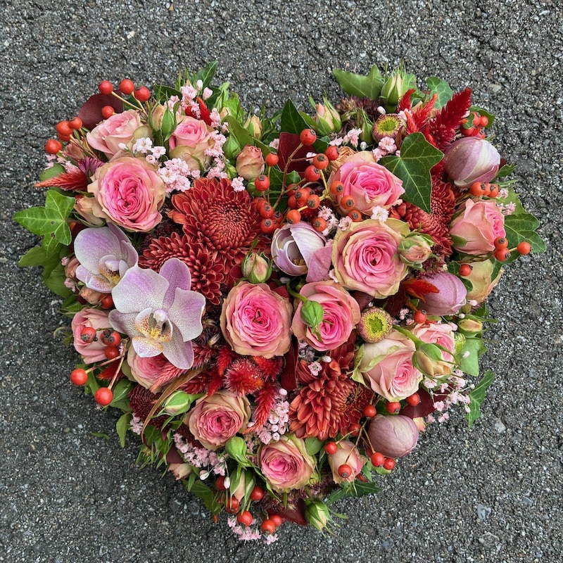 Herzförmiges Blumenarrangement aus Rosen, Orchideen und Beeren auf Asphalt, ideal für romantische Anlässe.