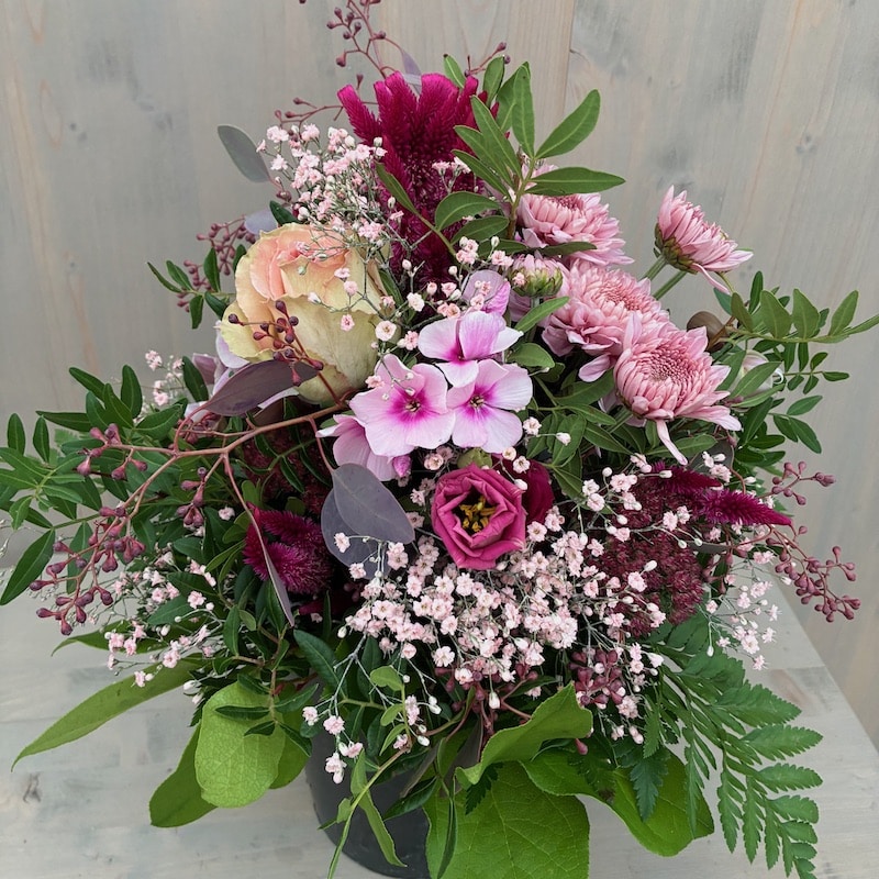 Ein bunter Blumenstrauß mit Rosen, Nelken und verschiedenen Blüten in einem schwarzen Topf, vor hellem Hintergrund.