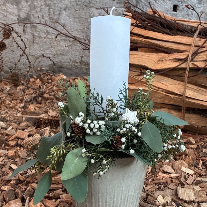 Weiße Kerze in rustikalem Adventsgesteck mit Tannenzweigen und Eukalyptus vor Holzstapeln.