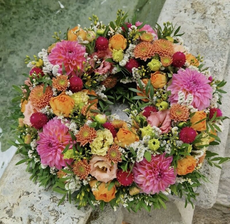 Bunter Blumenkranz mit rosa, orange und gelben Blüten, auf steinernem Hintergrund liegend. Ideal für festliche Dekorationen.