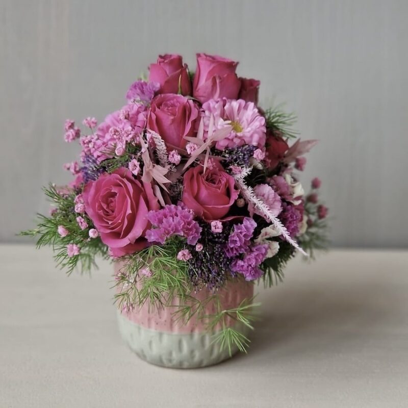 Blumenstrauß mit pinken Rosen und lila, rosa Blüten in rosa Vase, dekorativ arrangiert auf beigem Hintergrund.