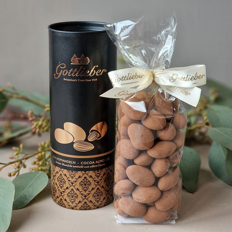Gottlieber Cacao-Mandeln, elegant verpackt, neben einer schwarzen Geschenkbox. Premium-Genuss seit 1928.